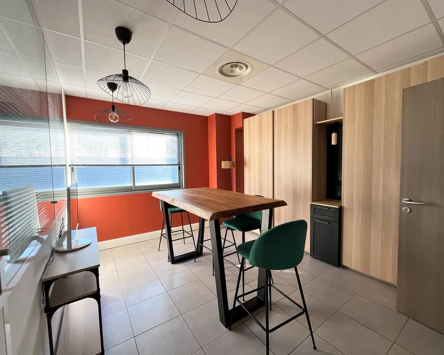 La salle de réunion de nos bureaux à Vendargues : mur terracotta, table haute en bois, tabourets verts et cloison vitrée.